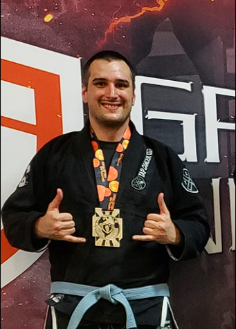 Scott Lee, a man in a black jiu-jitsu gi with a blue belt, smiling and showing a double shaka hand gesture while wearing a silver medal in front of a competition backdrop.